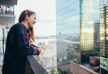 A young woman leaning against a balcony rail and looking at city buildings.