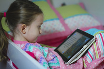 Girl in pajamas reading the Bible on an iPad and smiling. She's reading before bedtime.  Child doing devotions on a tablet. Reading a children's Bible.