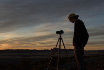 boy standing beside a camera on a tripod at sunset 
