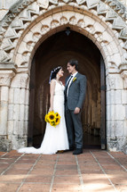 Bride with sunflower bouquet standing with groom on terra cotta tile in archway of church.