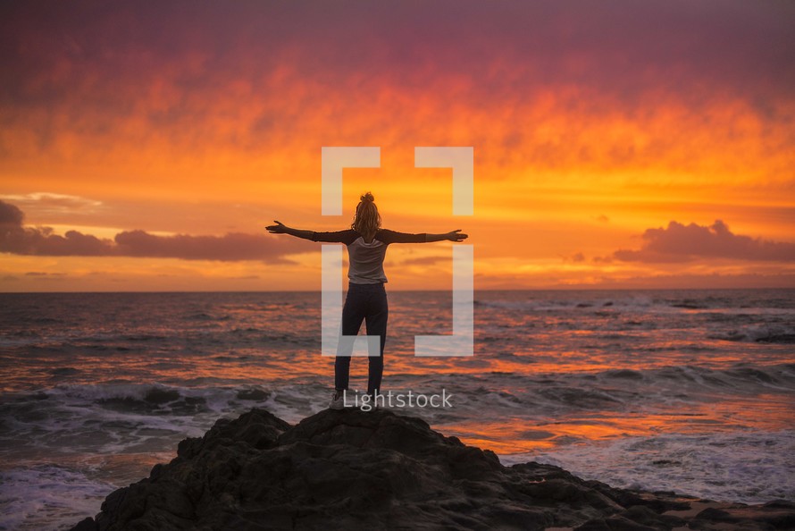 a woman standing on a shore at sunset with outstretched arms 