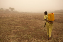 An Indian woman in yellow carries water on her back across a barren land.