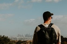 young man standing with a backpack looking out at a distant city 