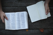 Hand on the open pages of a Bible on a picnic table with study notes.