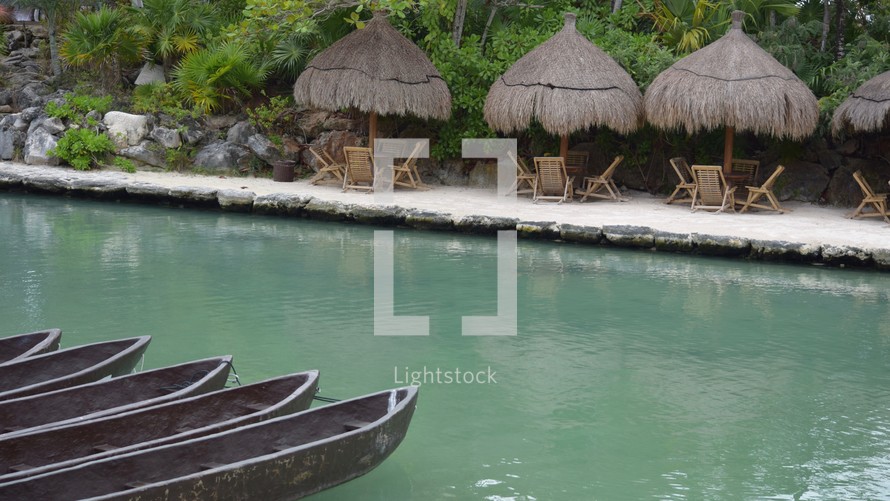 row of paddle boats across from straw umbrellas with table and chairs 