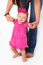 Mommy in jeans helping infant daughter in pink dress stand up and walk.