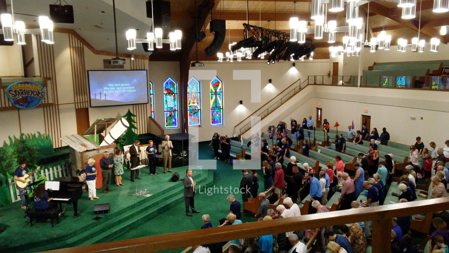 A Pastor leads his congregation in prayer during a traditional worship service surrounded by stained glass windows, a worship band and musicians during a Sunday morning time of worship and praise. 