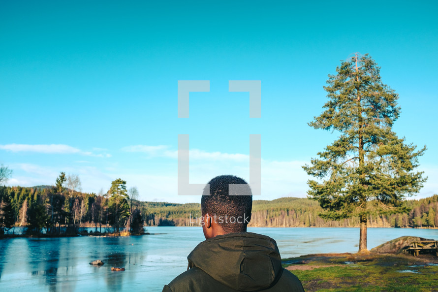 a man in a coat standing with his back to the camera looking out at a lake 