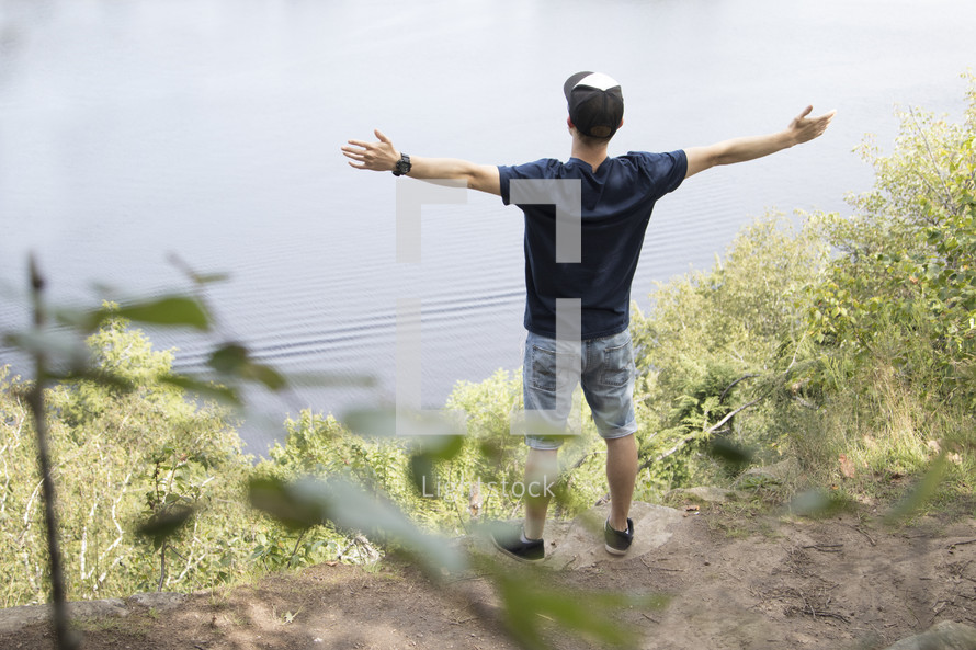 Teenager standing high on cliff overlooking lake with arms outstretched