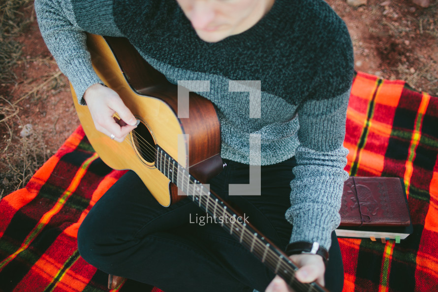 man with a guitar sitting on a plaid blanket 