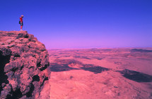 Man standing on a mountain cliff overlooking harsh desert terrain