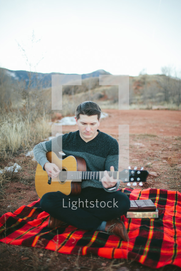 man sitting on a plaid blanket playing a guitar outdoors 