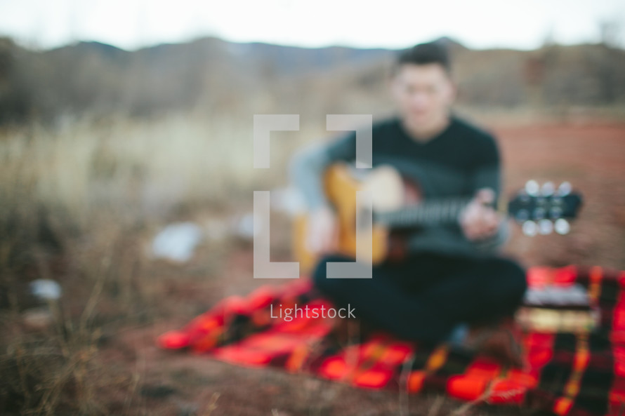 man sitting on a plaid blanket playing a guitar 