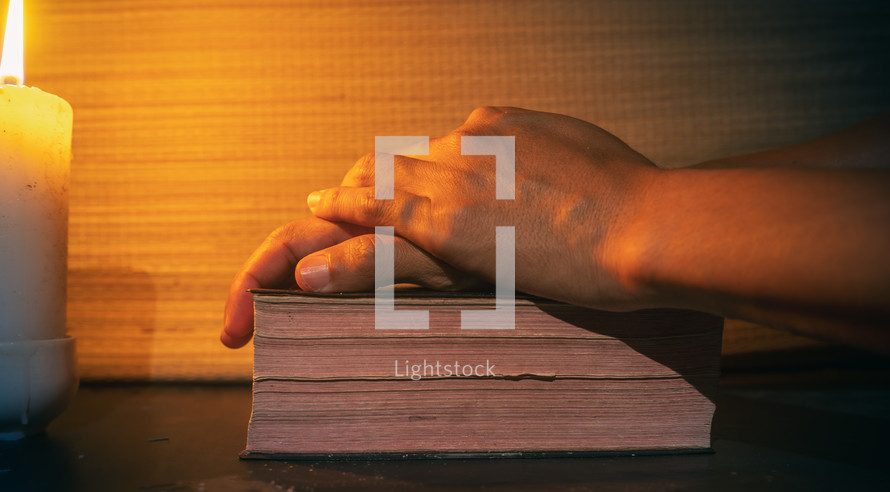 candle light with hands pray to bible on wooden table in the night time.