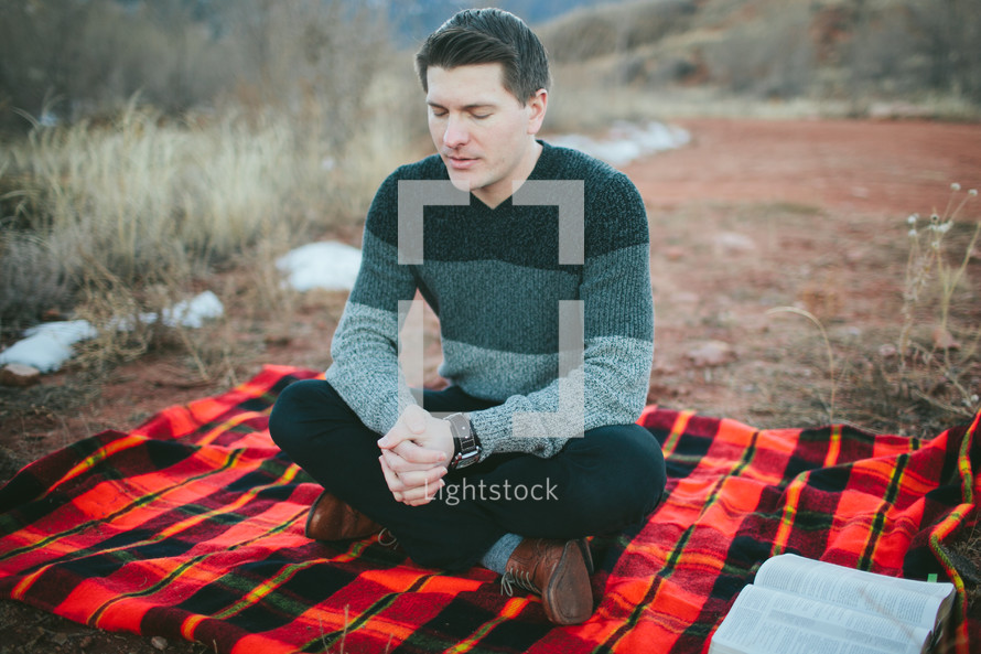 man with praying hands sitting on a plaid blanket 