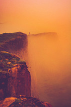 man standing on the edge of a cliff under an orange sky 