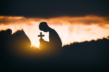 Young man praying and holding Cross In front of the stone at sunset background.