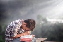 a boy praying outdoors over the pages of a Bible 