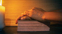 candle light with hands pray to bible on wooden table in the night time.