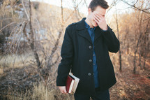 man hiding his face with his hands carrying a Bible outdoors 