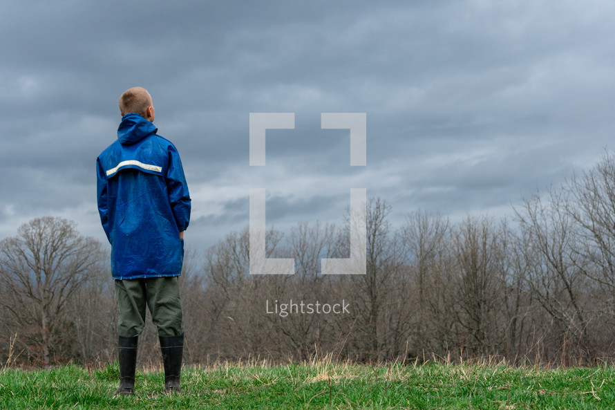 a boy in rain boots looking out a forest 