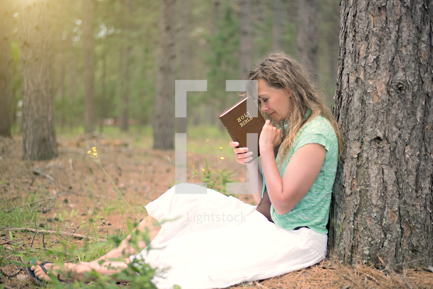 a woman sitting under a tree in a forest holding a Bible in prayer 