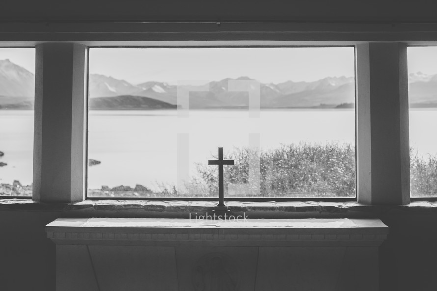 A cross in a window with a view of a lake and mountains.