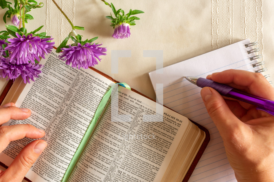 person taking notes and an opened Bible, pen, notepad, and flowers 