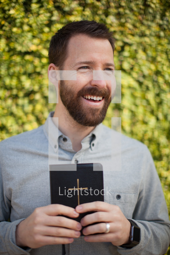 a smiling man with a beard holding a Bible outdoors 