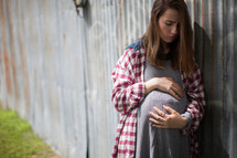 An anxious pregnant teenager holding her stomach and leaning against a wall.