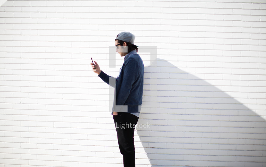 a man looking at his cellphone standing in front of a white wall 