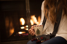 a woman sitting in front of a fireplace holding a mug of hot cocoa 