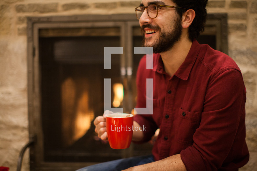 a man with a mug of hot cocoa sitting in front of a fireplace 