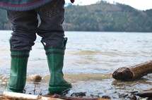 A child standing on the edge of a lake in rain boots.