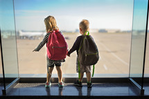 siblings holding hands looking out a window at an airport 