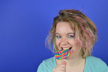 portrait of a young blond woman with colourful streaks in the hair biting into a big sweet multicolored lollipop and having fun looking into the camera