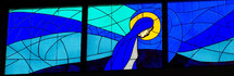A large stained glass window mural of Mary, the mother of Jesus, praying and surrounded by a halo and shades of blue and gold.