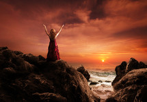 Woman standing on ocean rocks praising God at sunset.