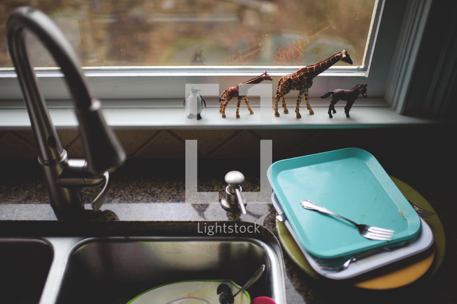 dirty dishes in a sink and plastic toy animals in a window sill 