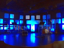 A sound stage complete with keyboards, drums, guitars all set up for a live praise and worship band to come and play before a live audience in a contemporary worship live church service. The background of the stage has a large cross and the stage is lit with blue lighting.