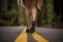 Legs of a woman walking along the center stripe of a highway through a forest.
