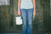 Woman standing in front of a wooden wall holding a paint can and brush