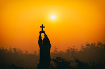 a male holding a cross at sunset against an orange sky