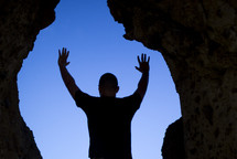 a man standing in the mouth of a cave with raised hands 