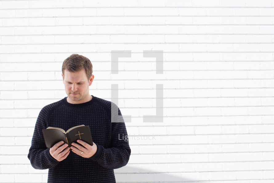 a man standing in front of a white wall reading a Bible 