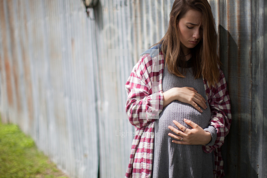An anxious pregnant teenager holding her stomach and leaning against a wall.