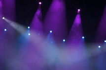 Purple and blue spotlights focused on concert performance stage through theatrical fog or smoke.