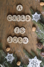 star ornaments, pine cones, snow, pine boughs and the words A CHILD IS BORN on wooden slices