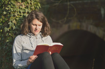 woman reading the bible while sitting next to a tree and a bridge in the evening light.