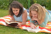 Young women smiling while reading in the bible together laying outside on a blanket in the grass on a sunny summer day. 
